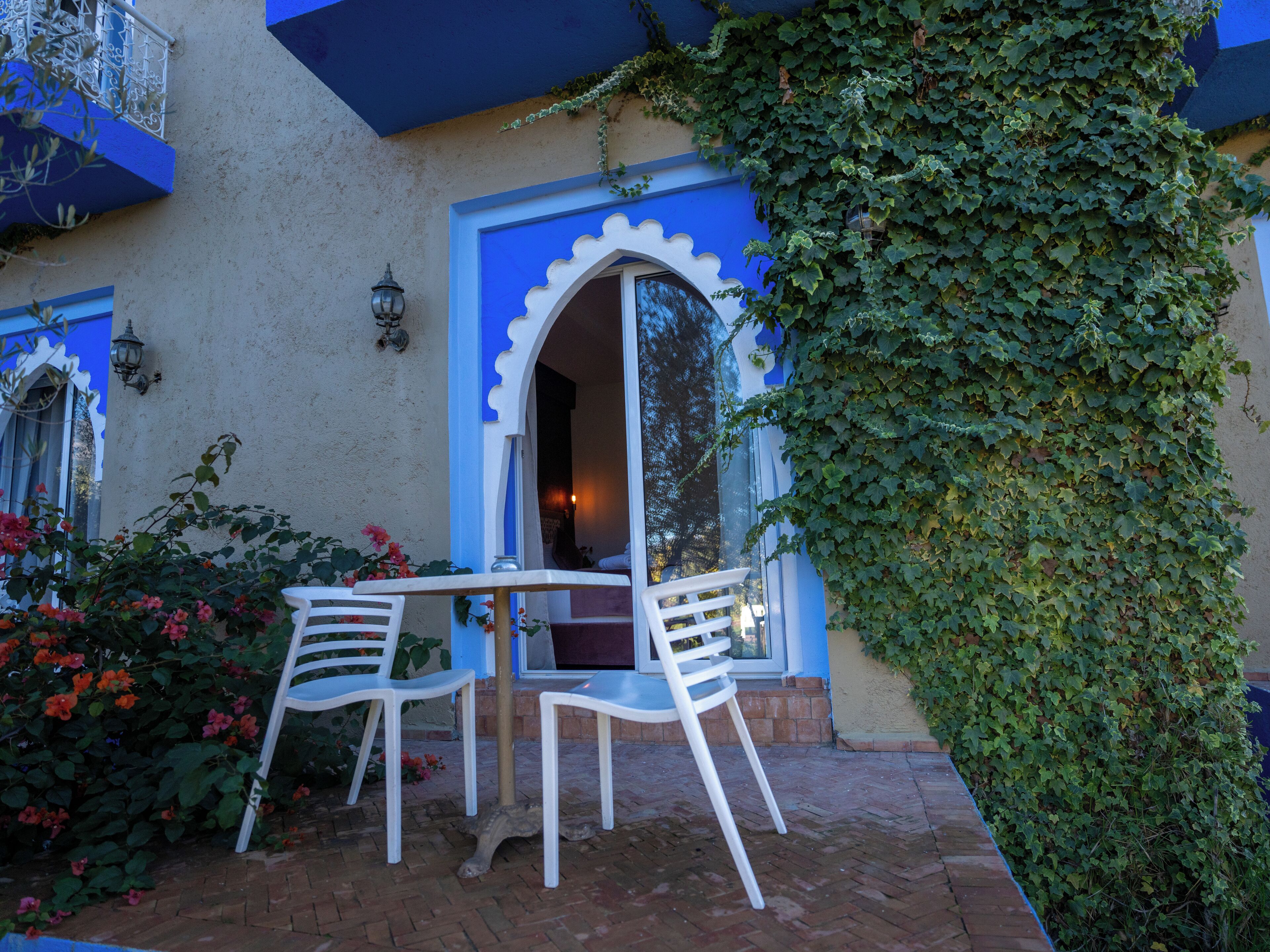 Un patio exterior con dos sillas blancas y una mesa, rodeado de piso de ladrillo y una pared cubierta de hiedra verde.