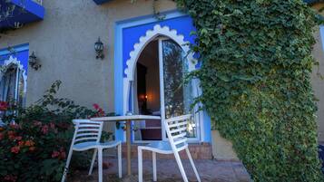Un patio exterior con dos sillas blancas y una mesa, rodeado de piso de ladrillo y una pared cubierta de hiedra verde.