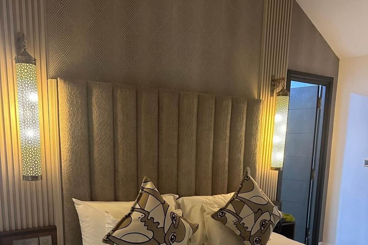 A hotel room with a large bed, two decorative pillows, a nightstand with a phone, and a wall-mounted lamp.