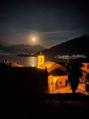 A night scene with a lit church and a moon in the sky, overlooking a body of water and distant hills.