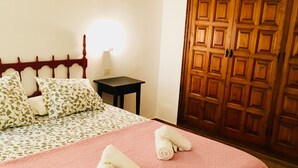 A neatly made bed with a pink blanket and two rolled towels, a wooden headboard, a bedside table with a lamp, and a wooden wardrobe.