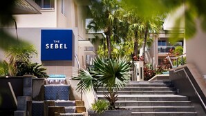 L’entrée d’un hôtel, avec une enseigne bleue portant l’inscription « THE SEBEL », une fontaine et un escalier menant au bâtiment.