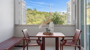 Un balcon avec une table et des chaises en bois, un banc et une vue sur la verdure et les bâtiments.