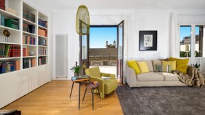 Een woonkamer met een boekenkast, een bank, een stoel en uitzicht op een gebouw door een geopende deur.