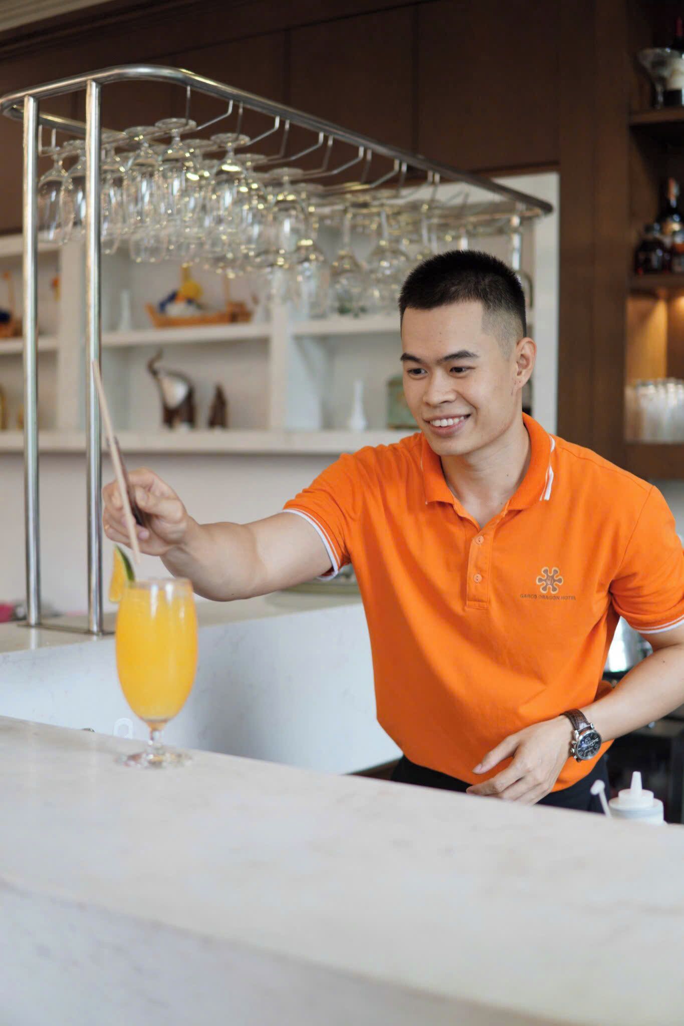 A person in an orange shirt preparing a drink at a bar with a wine rack in the background.