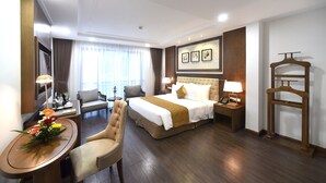 Quarto de hotel com cama, duas cadeiras, escrivaninha com luminária, mesa de cabeceira com luminária e guarda-roupa.