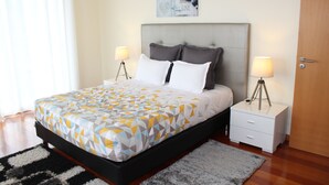 A neatly made bed with a geometric patterned bedspread, two bedside lamps, a small bedside table, and a framed picture above the bed. - Peaceful Escape Between Ocean and Mountains - Conceição House (Lombo Do Doutor)
