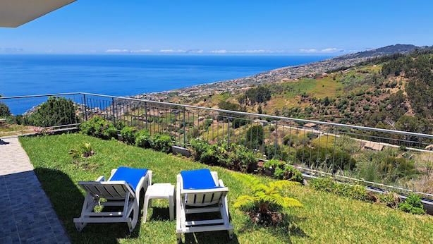 A terrace with two lounge chairs and a table, overlooking a coastal town and the sea. - Peaceful Escape Between Ocean and Mountains - Conceição House (Lombo Do Doutor)