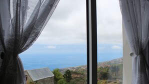 A window with sheer curtains, a view of a coastal landscape, and a distant town. - Peaceful Escape Between Ocean and Mountains - Conceição House (Lombo Do Doutor)