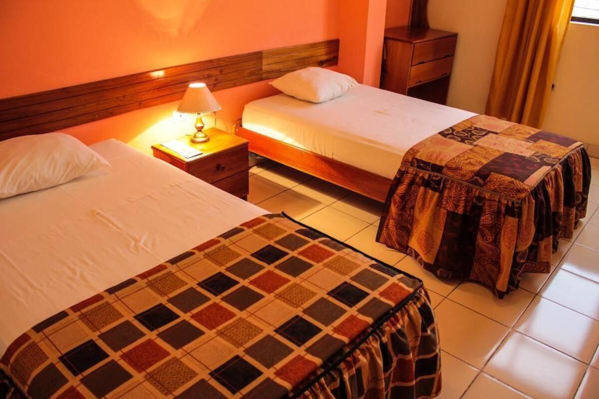 Quarto de hotel com duas camas, uma mesa de cabeceira com abajur, cabeceira de madeira e uma janela com cortinas.