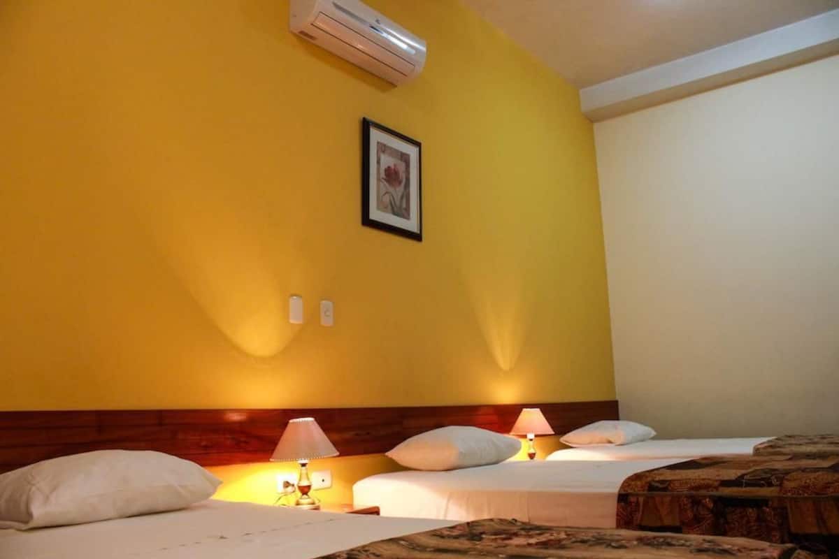 Quarto de hotel com duas camas, cabeceira de madeira, ar condicionado, quadro emoldurado na parede e abajures.