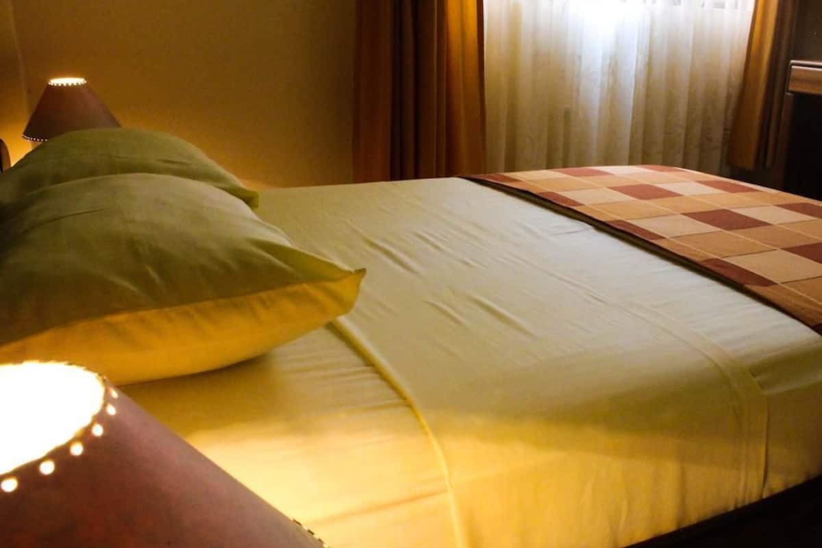 Uma cama com colcha xadrez e duas almofadas, um abajur ao lado e cortinas ao fundo.