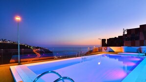 A swimming pool with a view of the ocean at dusk, surrounded by modern buildings.