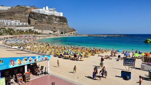 A beach with numerous umbrellas and sun loungers, a lifeguard tower, and a cliffside hotel in the background.