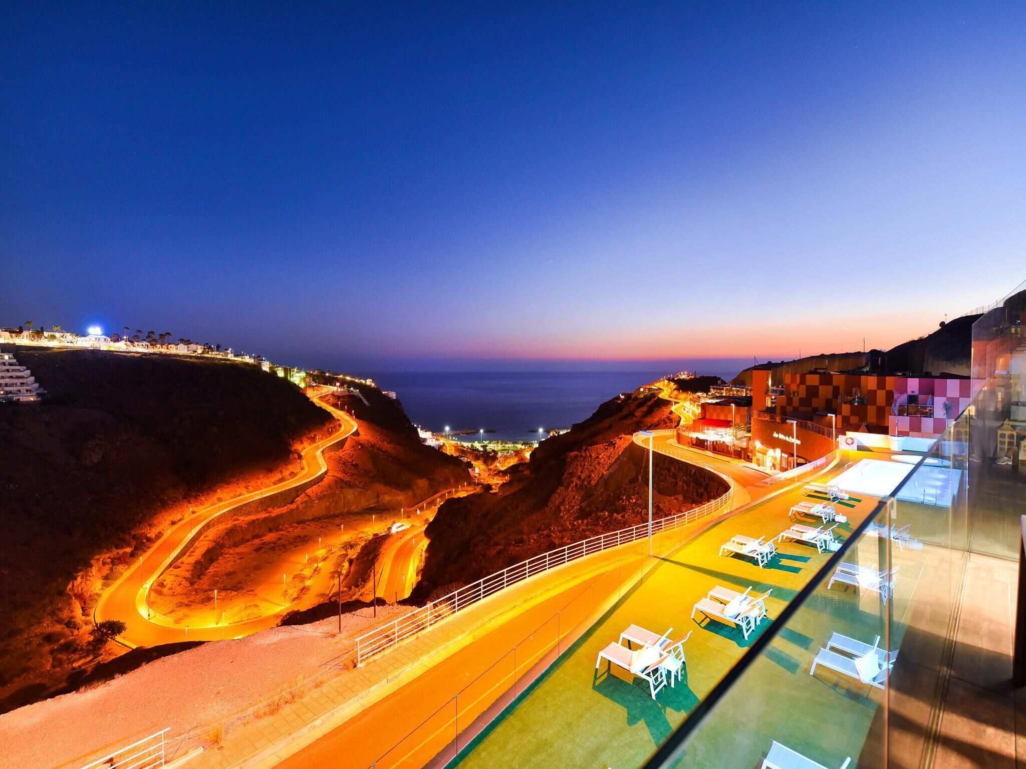 A coastal road with a winding path leading to a beach area with sun loungers and a pool.