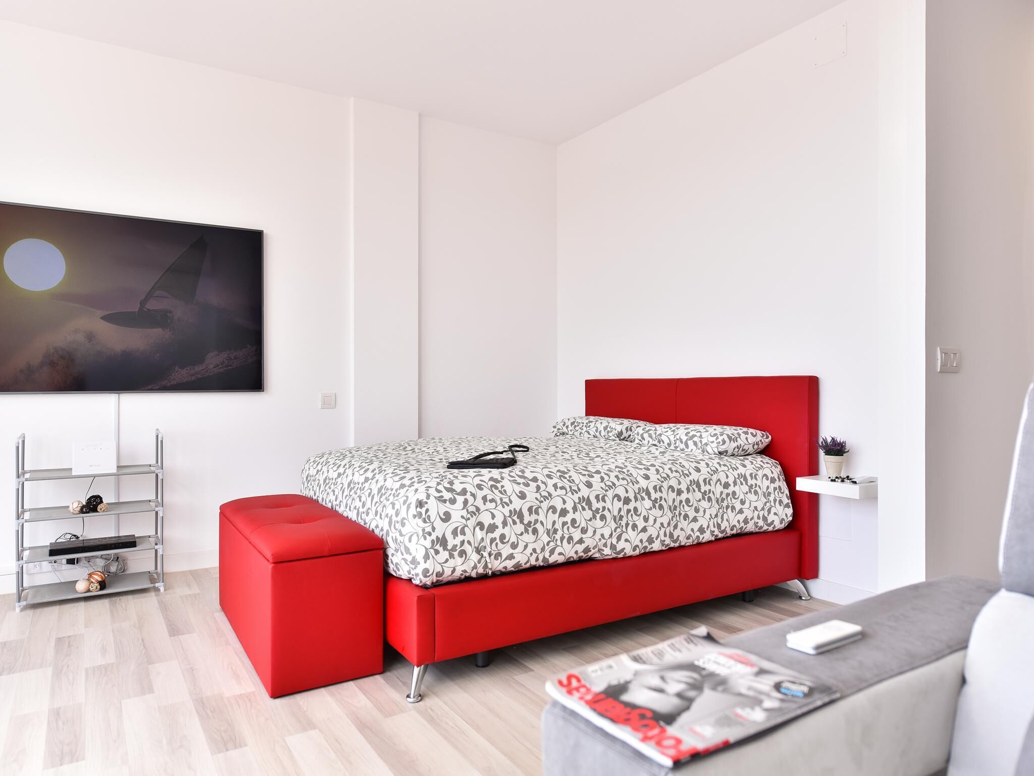 A modern bedroom with a red bed, a white wall, and a painting of a sailboat.