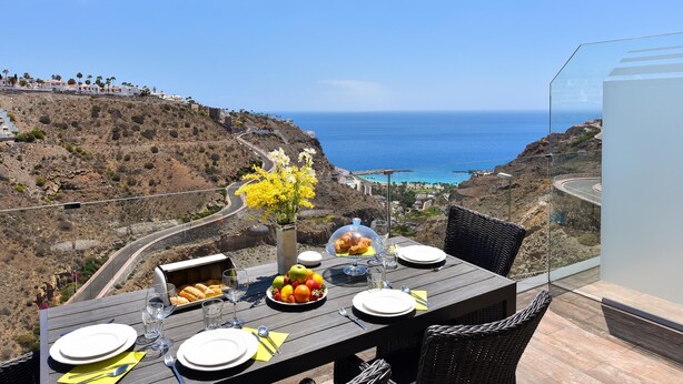 A table set for dining with a view of the ocean and a winding road.