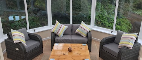 A conservatory with wicker furniture, a wooden coffee table, and large windows offering a view of greenery.