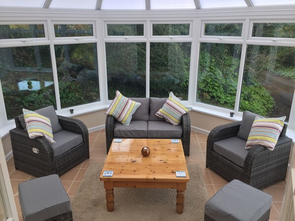 A conservatory with wicker furniture, a wooden coffee table, and large windows offering a view of greenery.