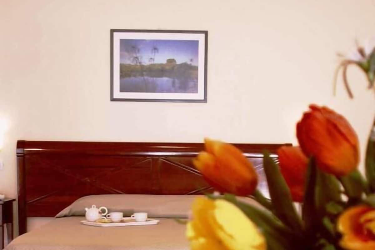 A hotel room with a bed, a bedside table with a glass of water, a tray with a teapot and cups, and a framed picture on the wall.