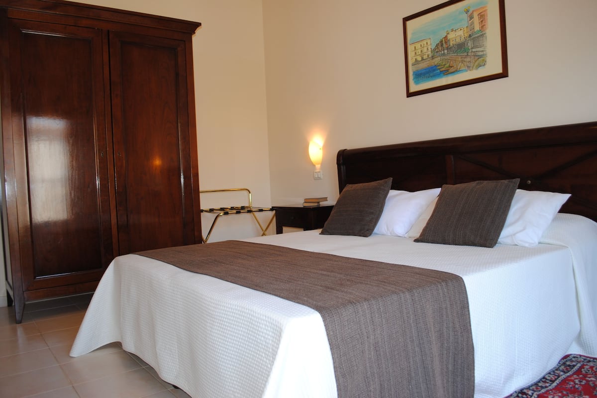 A neatly made bed with a white and brown bedspread, a wooden headboard, and a painting of a cityscape on the wall.