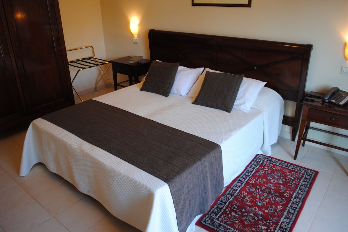 A neatly made bed with a dark headboard, a bedside table with a lamp, and a red patterned rug on the floor.