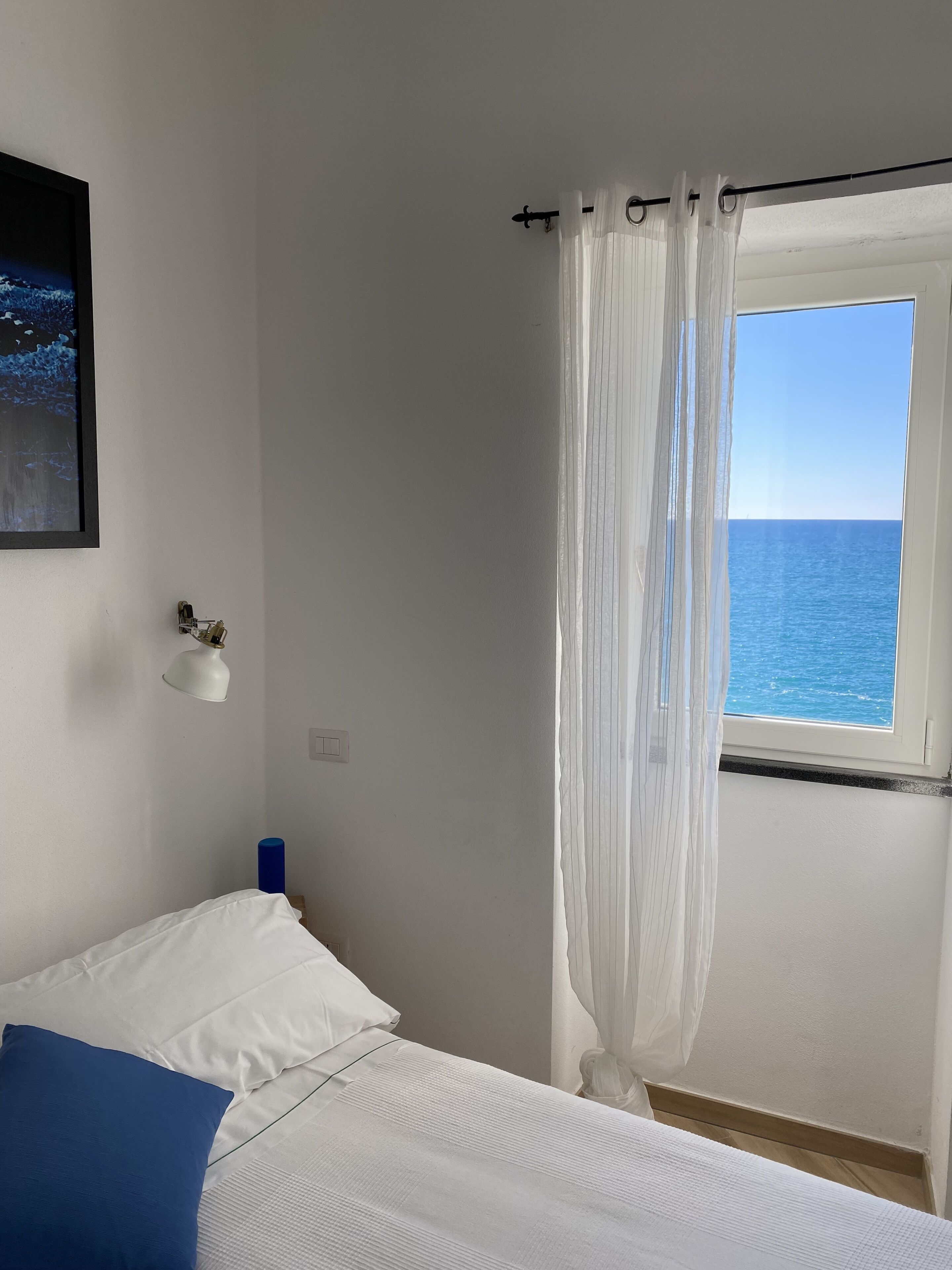 Ein Schlafzimmer mit einem Bett, einem Fenster mit transparenten Vorhängen, einem gerahmten Bild an der Wand und einem kleinen Regal mit Dekoration.