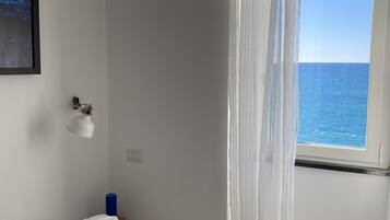 Ein Schlafzimmer mit einem Bett, einem Fenster mit transparenten Vorhängen, einem gerahmten Bild an der Wand und einem kleinen Regal mit Dekoration.