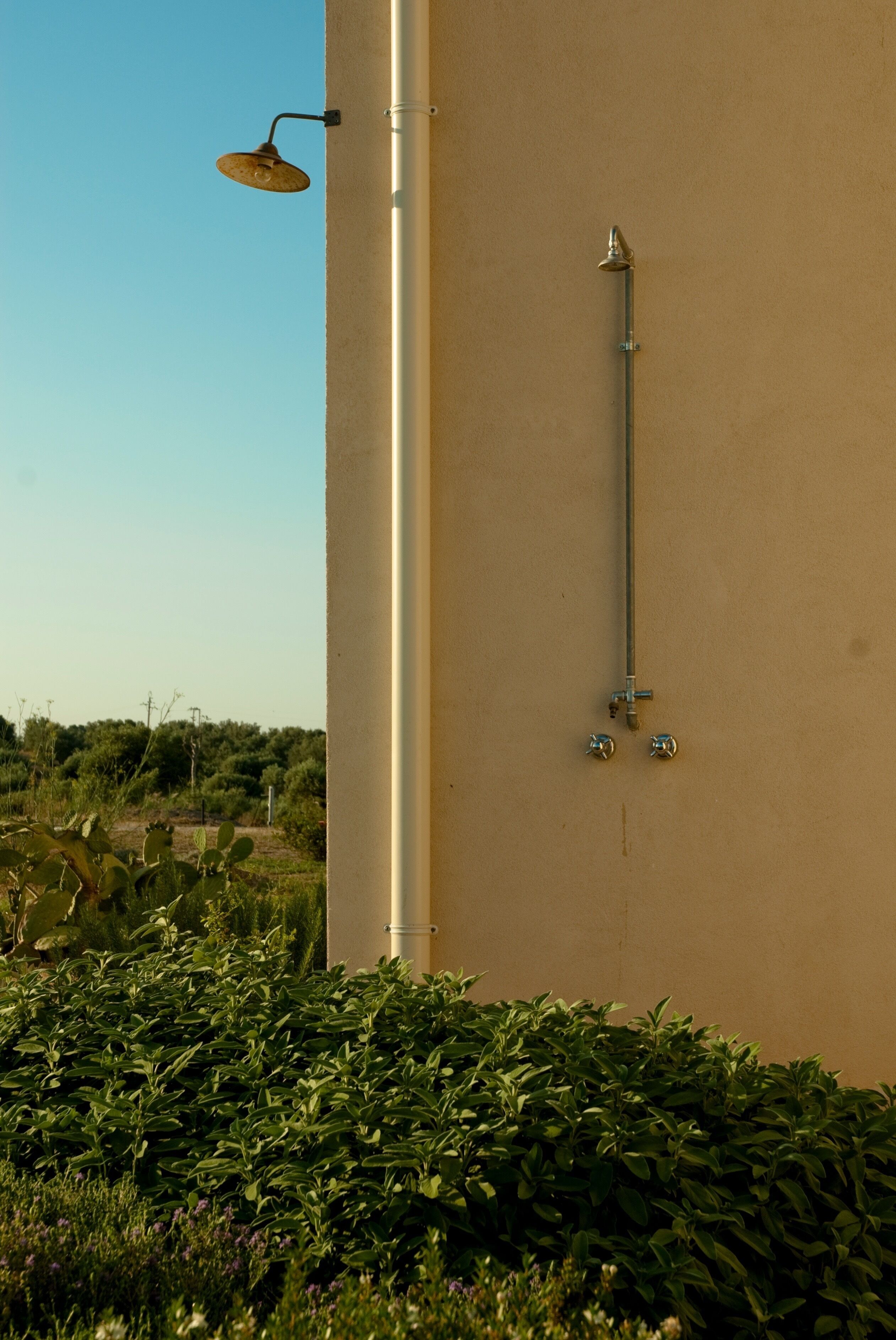 Un pommeau de douche fixé au mur et un luminaire sur un mur de couleur beige, avec un ciel dégagé et de la verdure au premier plan.