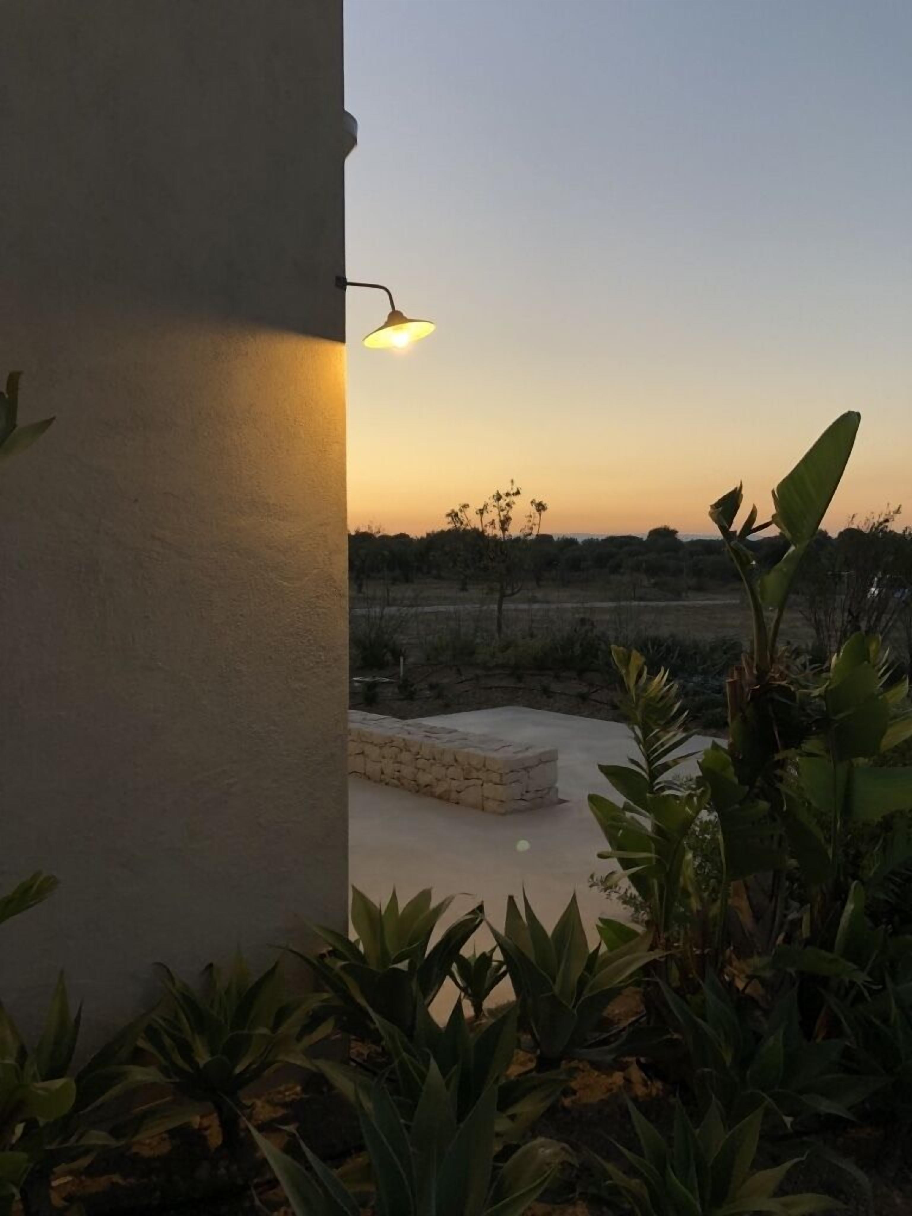 Un patio con un banco de piedra, rodeado de plantas, y vistas a un campo al atardecer.