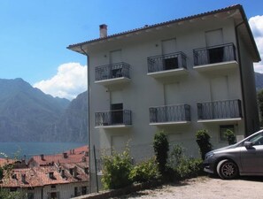 Un edificio de varios pisos con balcones, un coche estacionado al frente y montañas al fondo.