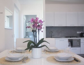 Une cuisine moderne avec des armoires blanches, un four encastré et une cuisinière à gaz. On y trouve une table à manger dressée avec de l’assiette blanche, des verres transparents et un vase rempli de fleurs violettes.