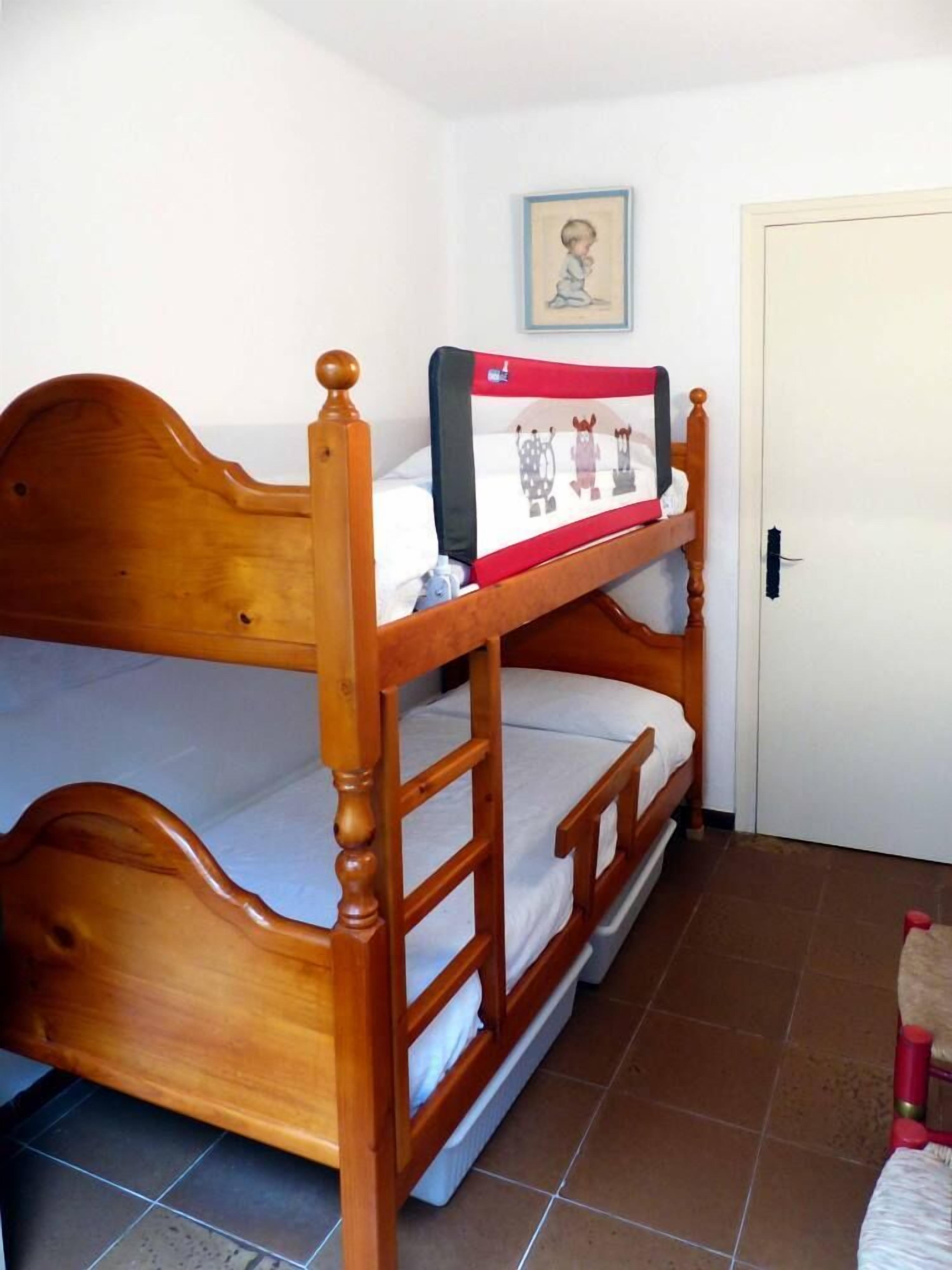 Un lit superposé avec une structure en bois et un matelas recouvert d’une housse rouge et blanche. Il y a une porte sur la droite et un tableau encadré accroché au mur au-dessus du lit.