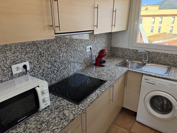 A compact kitchen with granite countertops, a built-in microwave, a red kettle, a sink, and a washing machine.