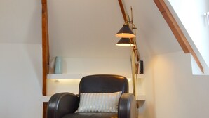A room with a sloped ceiling, a black leather chair with a patterned cushion, a floor lamp, and a small shelf with a vase.