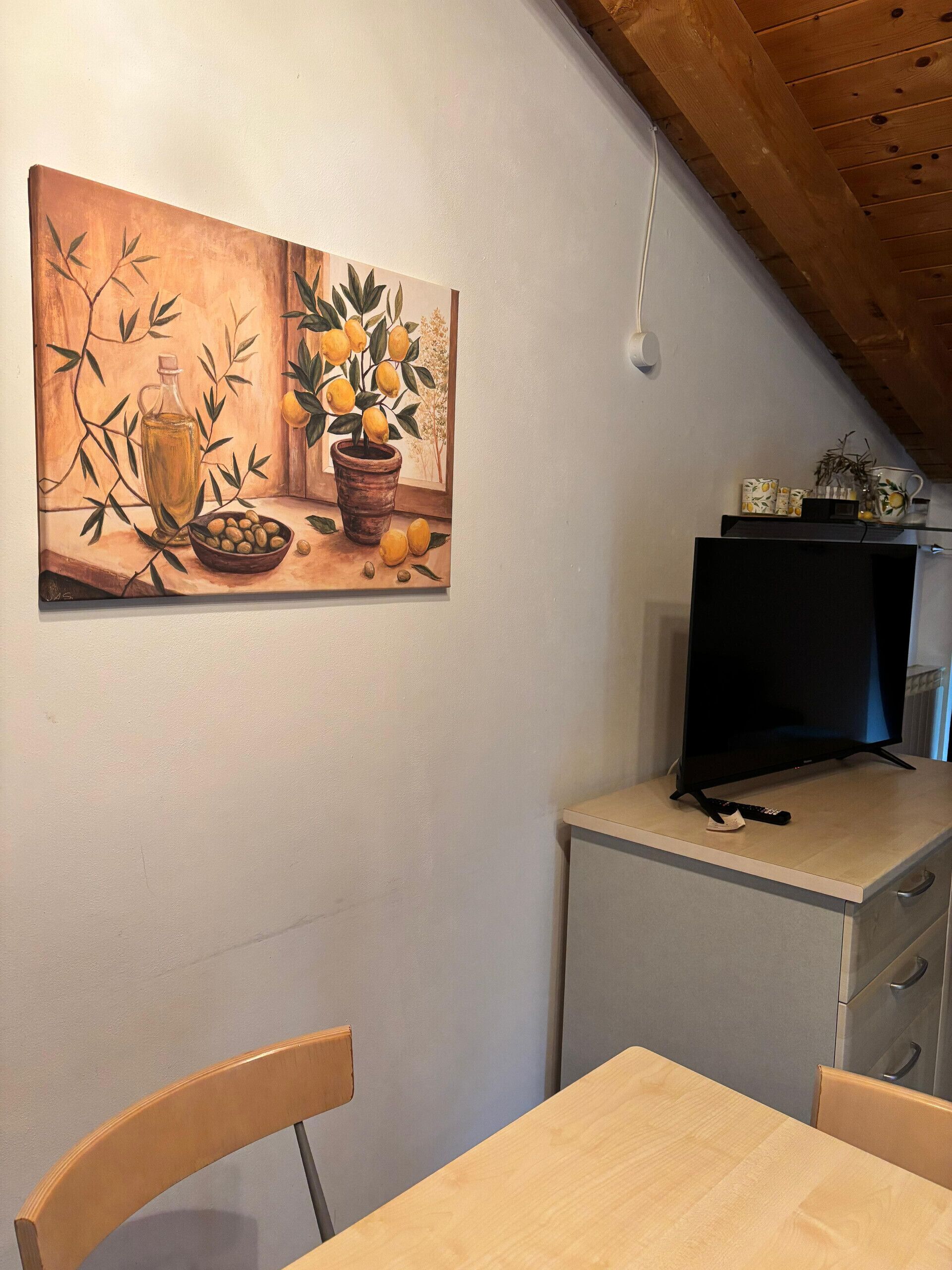 Un coin repas avec une table et des chaises, un tableau représentant des citrons et des olives, un téléviseur posé sur un meuble, et un plafond en bois.
