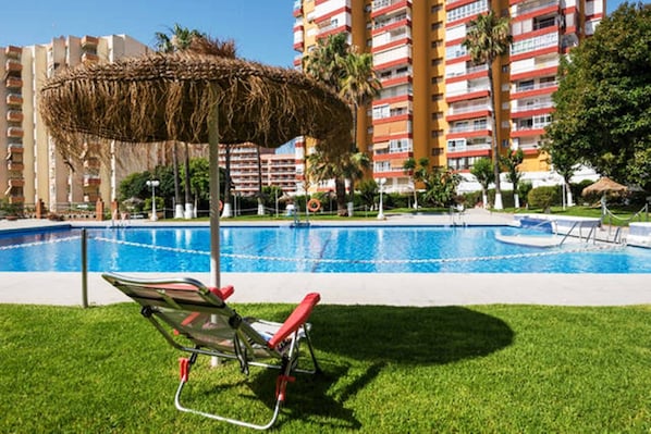 A swimming pool with a lounge chair and umbrella, surrounded by a grassy area and tall buildings in the background.
