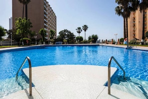 Una piscina con agua azul cristalina, rodeada de palmeras y un edificio alto al fondo.