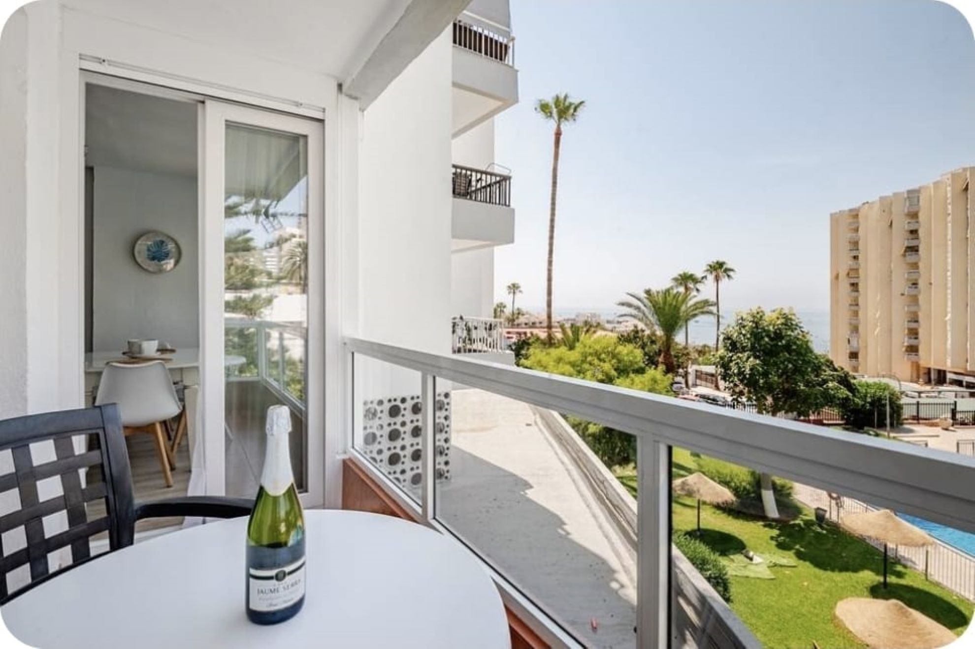 A balcony with a table and chairs, a bottle of wine, and a view of a pool and greenery.