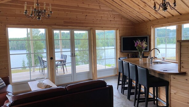 Interior de una cabaña con techo de madera, comedor con mesa y sillas, cocina con pileta y televisión, y ventanas amplias con vista a un lago.