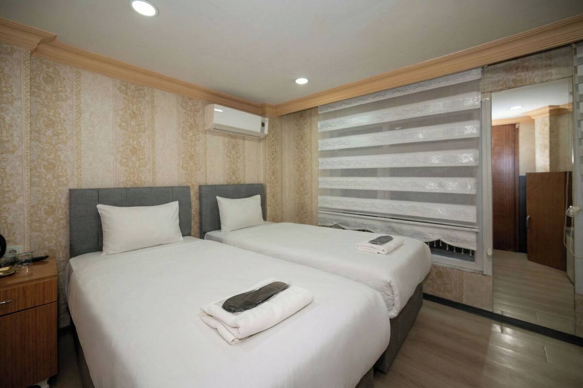 A hotel room with two beds, a wooden nightstand, a mirror, and a window with blinds.