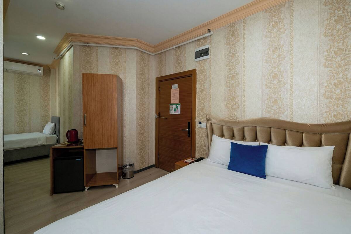 A hotel room with a bed, a wooden wardrobe, a small table with a red object, a white wall with a patterned design, and a door with a notice.