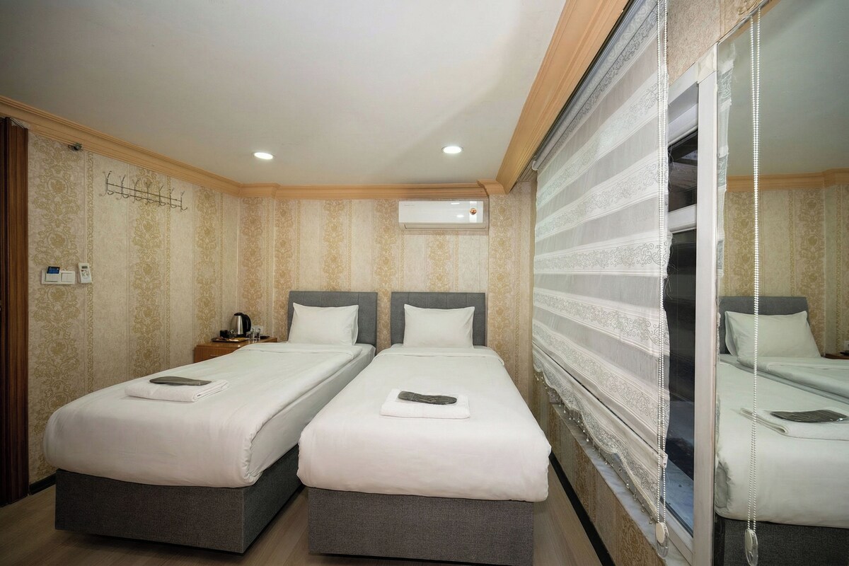 A hotel room with two beds, a bedside table with a lamp, an air conditioning unit, and a window with a curtain.