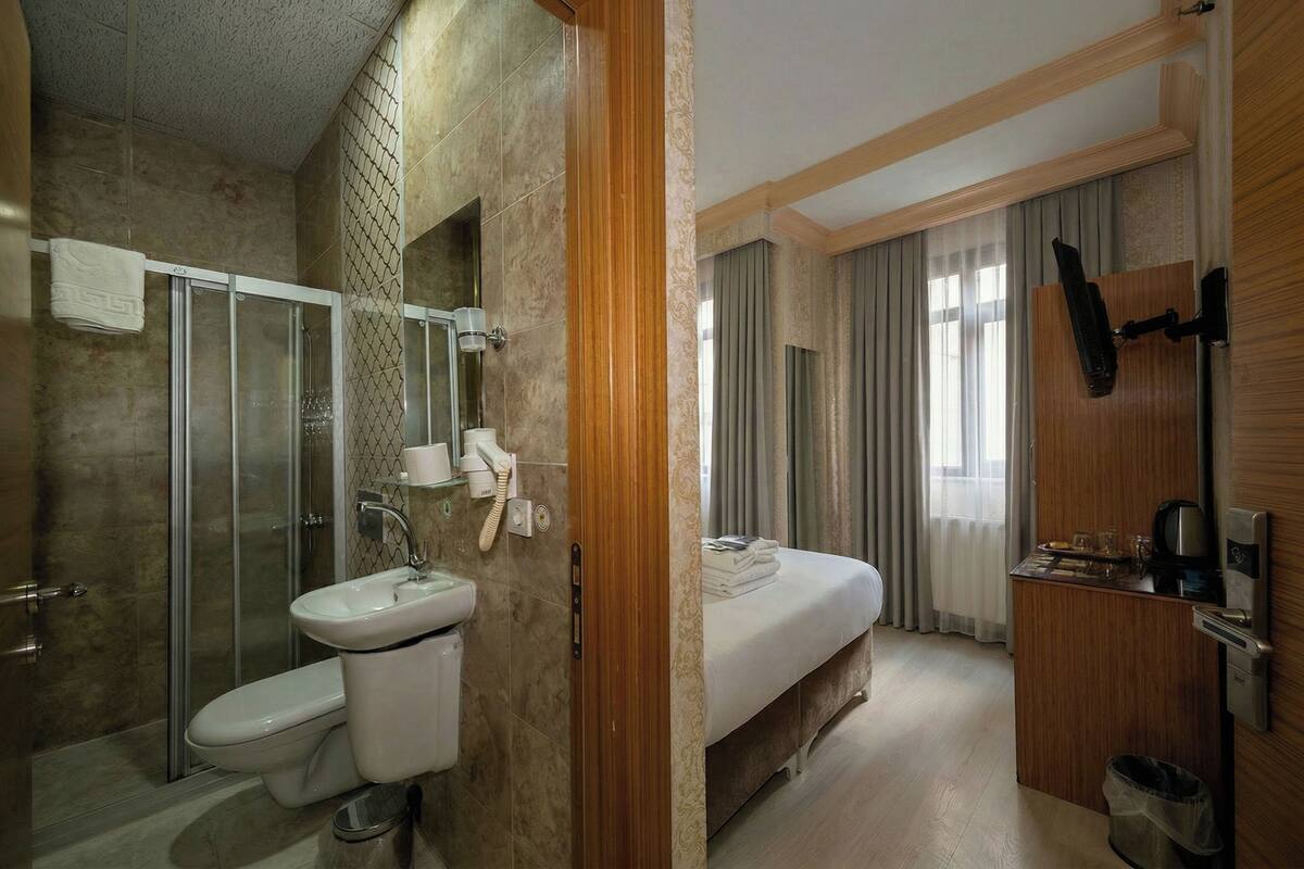 A hotel bathroom with a shower, toilet, sink, and a small bedside table with a coffee maker.