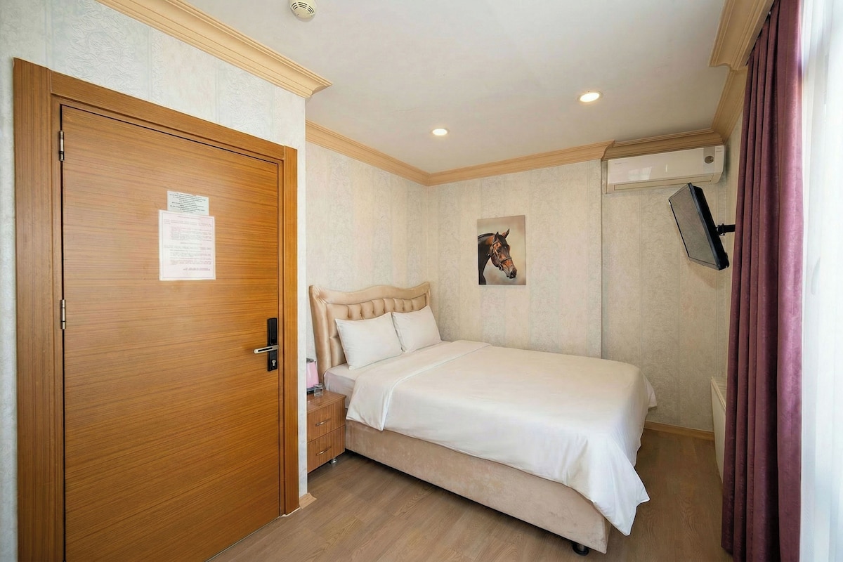 A bedroom with a wooden door, a bed with a headboard, a nightstand, a wall-mounted TV, and a ceiling light.