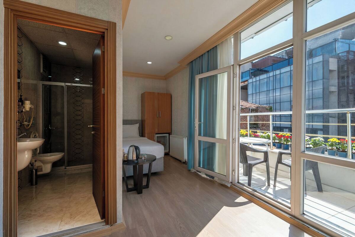 A hotel room with a bathroom, a bed, a small table, and a balcony with potted plants.