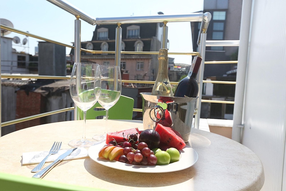 A table with a plate of fruit, two wine glasses, and a bottle opener on a balcony with a view of buildings.