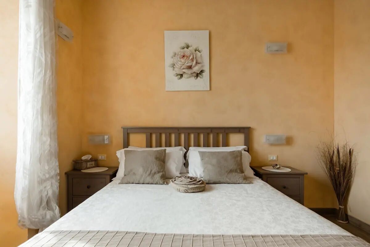 Une chambre à coucher comprenant un lit, deux tables de chevet, une petite table, une lampe fixée au mur, un coussin décoratif et un vase contenant des plantes séchées.