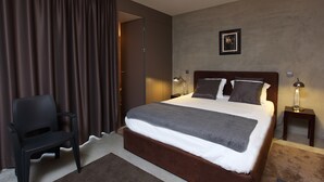 A neatly made bed with a grey blanket, a wooden headboard, and two bedside lamps. A black chair is positioned to the left of the bed.