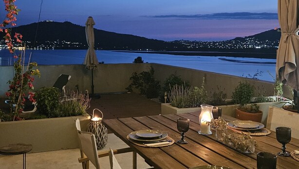 An outdoor dining area with a view of the sea and city lights at dusk.
